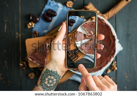 Woman hands make smartphone photo of raw vegan dessert. Chocolate sweet tart with dates (cake or pie). Phone photography of lunch or dinner for social networks. Top view.Vegetarian healthy food Royalty-Free Stock Photo #1067863802