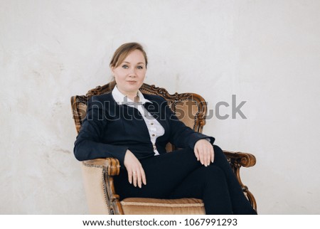 young beautiful blonde girl with blue eyes in a business suit sits in a vintage chair on a light background and looks around