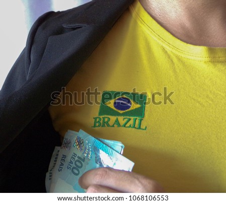 A person wearing brazilian shirt, hiding real money ( Brazilian money) into pocket. Financial help. Bribe and corruption. Giving money. Reference to Brazil corruption. Football corruption.