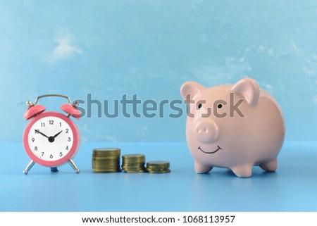 A small pink alarm clock and a piggy bank stand on a blue table. Between them are three stacks of coins of different heights. Beautiful bright photo. The symbol of the growth of money.