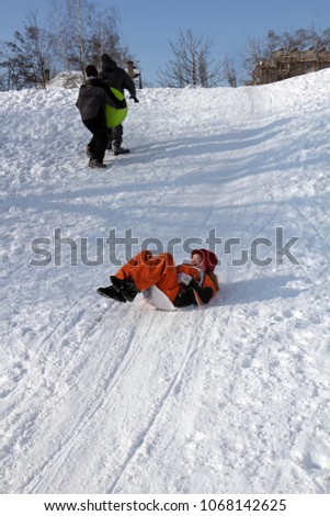 a little girl in an orange overalls slides down a snow hill