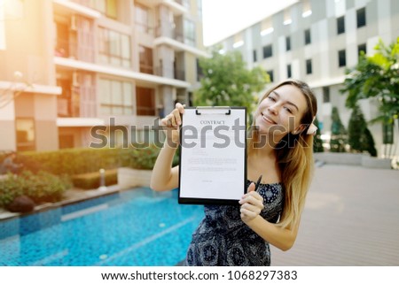 young pretty woman offer to sign contract on background of new apartment building. concept of long term rent of car, condo or traveling. Front used with Open Font License
