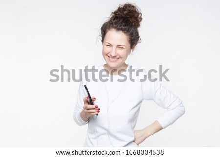 Happy young Caucasian female model with broad smile, laughs joyfully as hears anecdotes online in headphones, connected to modern smart phone, publishes internet post on website, poses at studio wall.