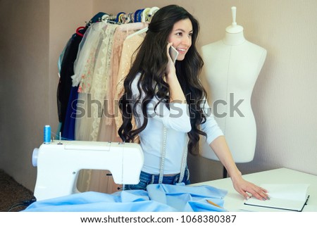 portrait of beautiful seamstress brunette with long hair talking on the phone . tailor creates a collection of outfits . young woman fashion designer dressmaker discusses outfit with customer by phone