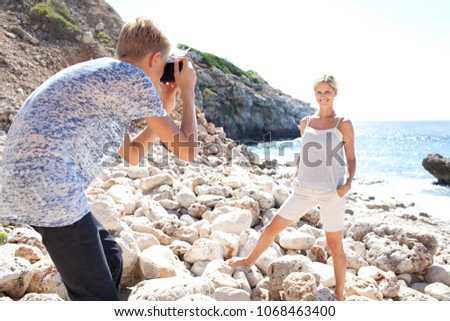 Mother and son on rocky beach holiday together with son using photographic camera taking pictures of mum on sunny day, outdoors. Family fun artistic activities, travel leisure recreation lifestyle.
