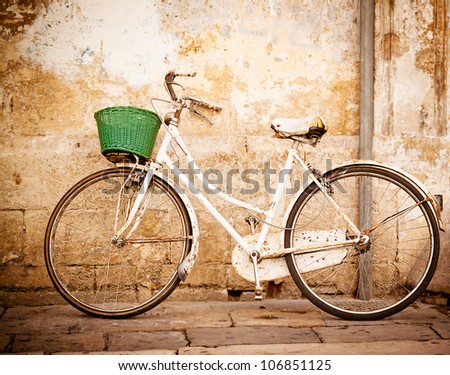 An old, rusty white bicycle with a basket leaning against a grungy wall in Italy. Royalty-Free Stock Photo #106851125