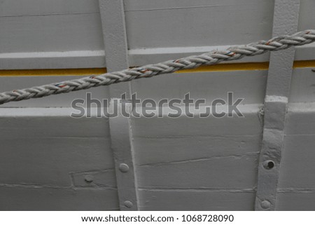 Close up outdoor view of part of a white ancient wooden boat. Large oblique rope in front of the hull made of planks. Yellow decorative painted line. Abstract picture of the detail of on old vessel.