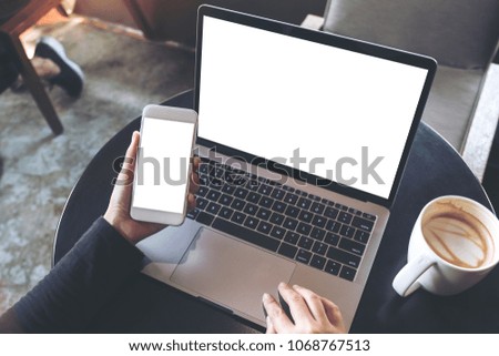 Top view mockup image of hands holding blank mobile phone while using laptop with blank white desktop screen on table 