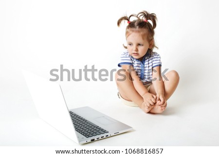 Attractive small female child sits in front of opened laptop, watches intersting colourful cartoons, has focused expression in laptop computer isolated over white background. Childhood and technology