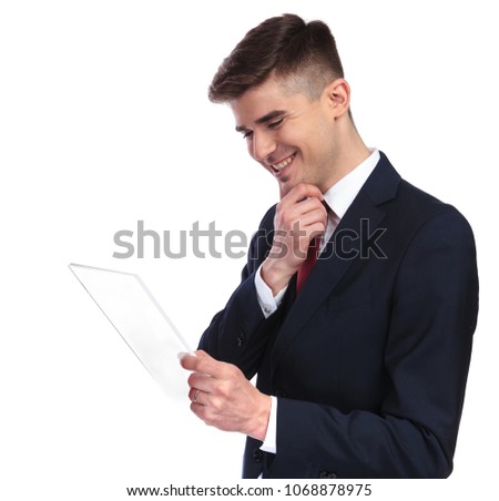 side view of curious businessman laughing while reading something on futuristic gadget. He wears a navy suit and a red tie while standing on white background, portrait picture.