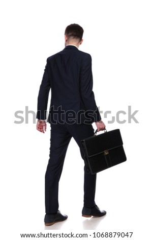 back view of businessman with suitcase walking while looking down to side at something. He wears a navy coloured suit, full body picture