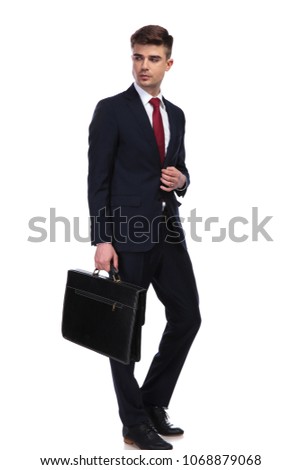 Businessman looks to side while holding his briefcase, standing on white background. He unbuttons his navy suit jacket; full body picture.