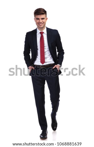 happy relaxed businessman wearing a navy suit and a red tie, walking with hands in pockets and smiling while standing on white background, full body picture