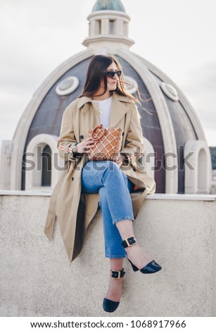 fashion details. fashion blogger posing on a rooftop in her elegant beige trench coat with a side stripe, denim jeans, ankle strap shoes. holding a leather net tote handbag.