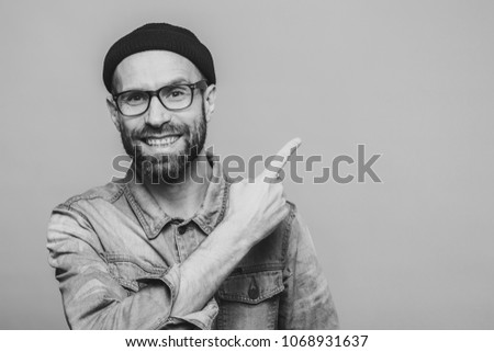 Horizontal shot of cheerful bearded man with happy expression, dressed in fashionable clothing, wears glasses, hat and denim shirt, indicated with fore finger at blank copy space for your advertisment