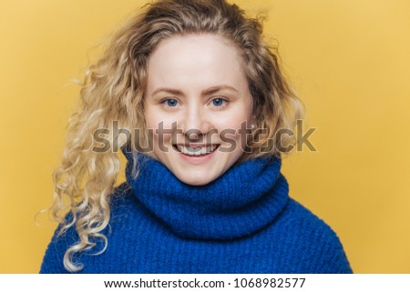 Happy delighted woman with light crisp hair, wears warm blue sweater, has broad smile and white teeth being satisfied after successful day, isolated over yellow background. Positive emotions