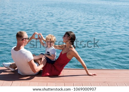 Mother and father making heart or love gesture with hands near their child. Happy family spend time together, sea background. Parents with son sit on seafront, rear view. Family vacation concept.