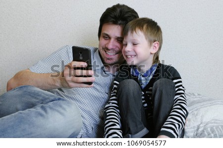 Dad and son are watching something in the mobile phone together and smiling.
