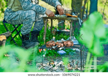 The meat is fried on an open fire with a large piece of skewer. Background-male tourist in camouflage sets the table. Field kitchen.