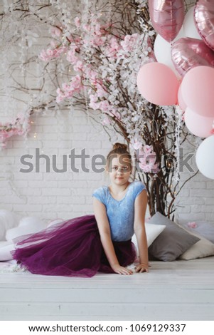 A young ballerina in blue clothes and a purple skirt with pink pointes sits on a white bed with pillows of branches of trees and pink balloons.