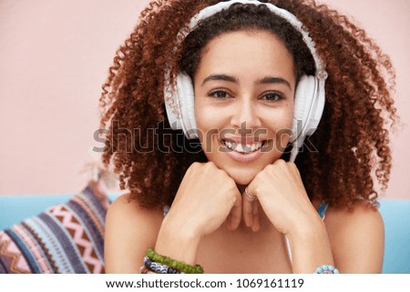 Close up shot of beautiful delighted young African woman has broad smile, enjoys favourite melody in headphones or listens online training lecture, while waits for friend in cozy calm cafeteria