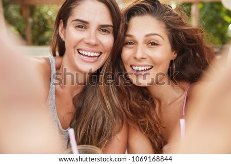 Portrait of happy women have broad smiles and satified expressions, stand close to each other as pose for selfie, going to share photos in social networks or publishing in own blog, rest in cafe