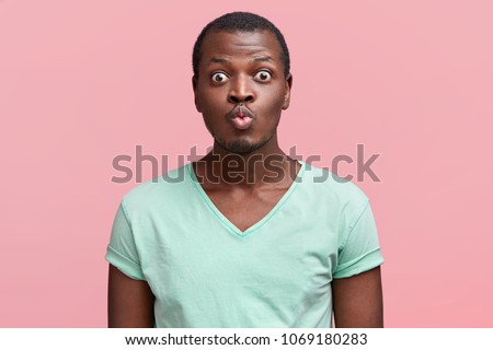 Portrait of positive delighted comic African American male with bugged eyes and rounded lips, dressed in casual t shirt, isolated over pink studio background. Handsome young man going to kiss you