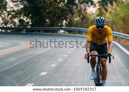 Close up on Asian man wearing a yellow cycling jersey, who's riding a road bike up high on hill in the morning. Under morning sunshine with determination on his face. Royalty-Free Stock Photo #1069182317