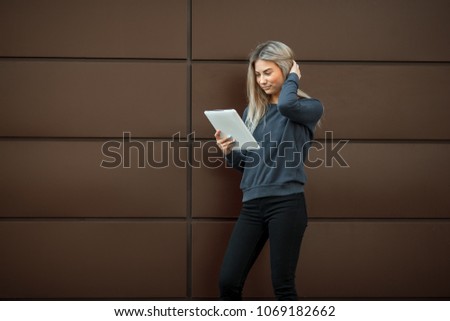 A young pretty girl is surfing on the internet on a tablet in the street on a brown modern background.