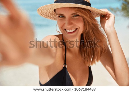 People, lifestyle, happiness and summer time concept. Lovely young smiling woman with cheerful expression poses for making selfie against azure sea background, happy to have good unforgettable rest