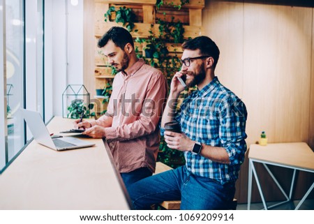 Young smiling hipster guy making telephone call while his colleague searching information in networks on laptop computer,male students ignoring real communication on coffee break using gadgets