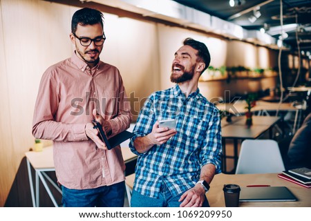 Young smiling hipster guy laughing at funny story telling by colleague on break in office, emotional cheerful man joking on friend networking on smartphone getting his funny pictures of email