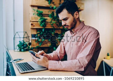 Serious young male freelancer disappointed with bad news while checking mail on smartphone sitting in coworking space,stressed hipster guy angry about reading banking balance via app on cellphone