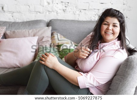 People, happiness, fun, joy and body positivity concept. Picture of cheerful joyful young Caucasian brunette female in leggings and plus size pink shirt, laughing, showing her perfect white teeth