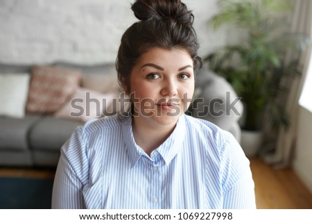 Beauty and body positivity concept. Picture of happy attractive young brunette plus size woman with hair bun and beautiful features posing indoors against gray couch and plant pot background