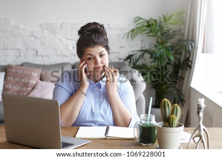 Picture of successful young overweight self employed woman in formal shirt working remotely from home office, speaking on mobile phone to customer, writing down important information in copybook