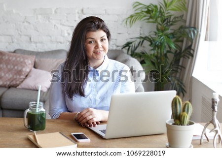 Picture of confident friendly looking young businesswoman with curvy body working on laptop pc at home office, sitting at her workplace, wearing stylish blue shirt, smiling broadly at camera