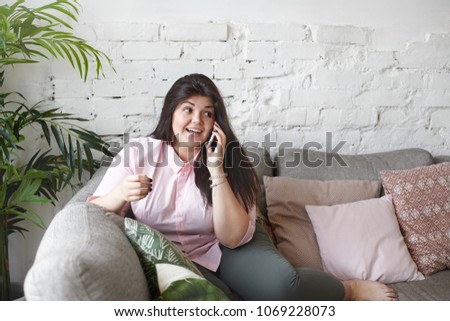 People, lifestyle and modern technologies concept. Picture of gorgeous chubby young woman talking on mobile phone, inviting friends to her place for party, smiling excitedly, sitting on sofa alone