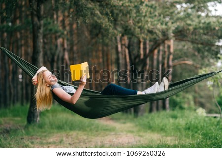 Young beautiful blonde girl in hammock reading e-book. Pretty woman leisure lifestyle portrait at nature ountdoor. Female relax in forest. Adorable teen expressive face. Tourism in summer camp. Hobby