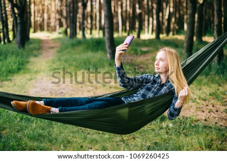 Young beautiful blonde girl in hammock and making selfie. Pretty woman leisure lifestyle portrait at nature ountdoor. Female relax in forest. Adorable teen face expression. Tourism in summer camp
