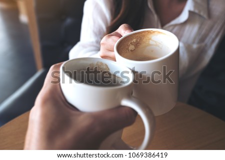 Close up image of a man and a woman clinking white coffee mugs in cafe
