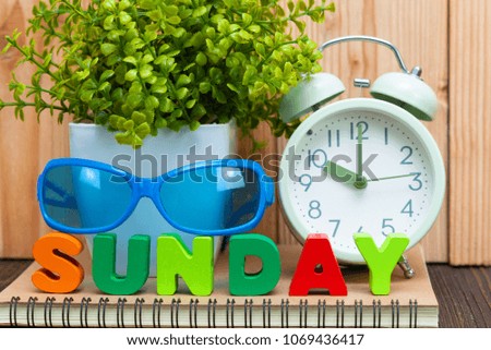 SUNDAY letters text and notebook paper, alarm clock and little decoration tree in white vase on wooden background, hello Sunday weekend concept idea