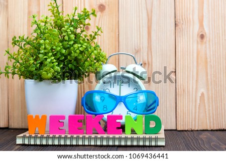 WEEKEND letters text and notebook paper, alarm clock and little decoration tree in white vase on wooden background, hello weekend vacation concept idea.