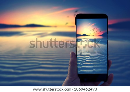 Hand holding Smartphone and taking photo of beautiful sunrise at beach