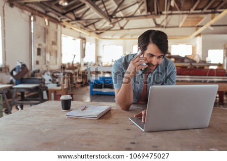 Smiling young craftsman standing at a workbench in his large workshop working online with a laptop and discussing designs with a customer on a cellphone Royalty-Free Stock Photo #1069475027