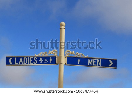 White wrought iron sign pole with blue signs indicating ladies’ and mens’ toilets set against blue sky