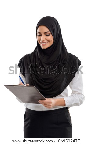 Happy female Muslim office worker. Woman writing on clipboard and giving a wide smile. Good job, employee satisfaction.