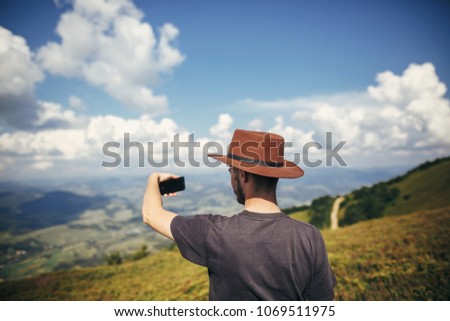 stylish traveler man in hat holding phone, walking on top of sunny mountains and sky. travel and wanderlust concept. space for text. happy hipster traveling and taking photo.