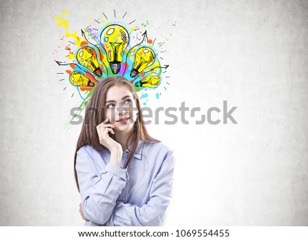 Dreamy young woman with brown hair wearing a blue shirt and business suit pants is looking upwards and thinking. A concretre wall background with many lightbulbs. Creativity concept. Mock up