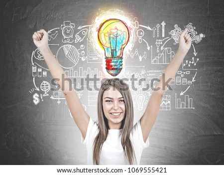 Smiling young woman with brown hair wearing a white t shirt is standing with her hands in the air and looking at the viewer. A blackboard background with a business idea sketch